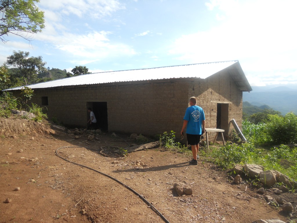 Two people stand outside an unfinished brick building with a metal roof on a dirt and rocky hillside, surrounded by greenery and mountains under a partly cloudy sky.