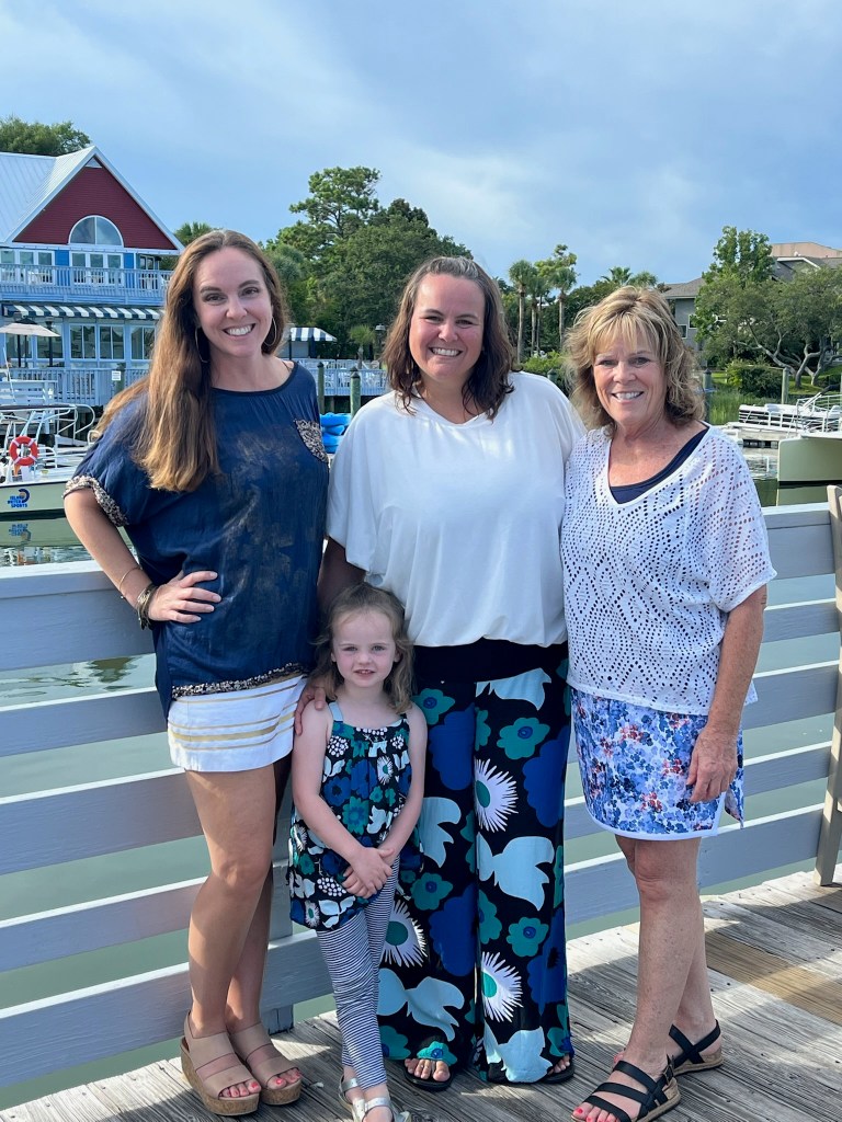 Four people, including a young girl, stand and smile on a wooden dock with water, boats, and a building with a red roof in the background.