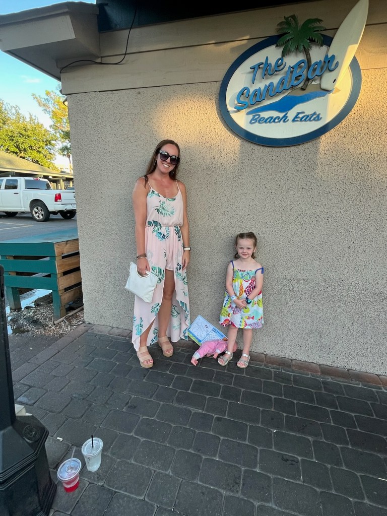 An adult and a young girl stand smiling outside The Sandbar Beach Eats restaurant; drinks and a drawing are on the ground nearby.