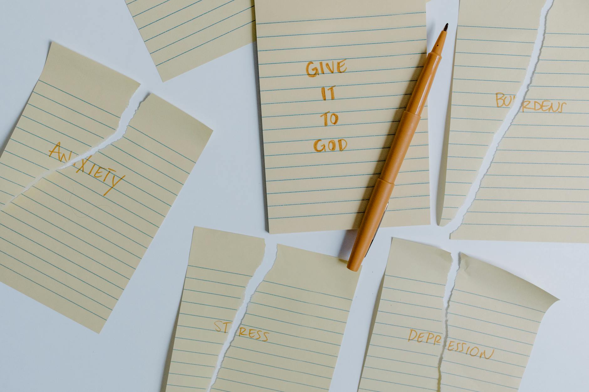 Six lined yellow papers are arranged on a white surface. Four are torn and labeled “anxiety,” “stress,” “burdens,” and “depression.” A whole one says “Give it to God” with a pen on top.